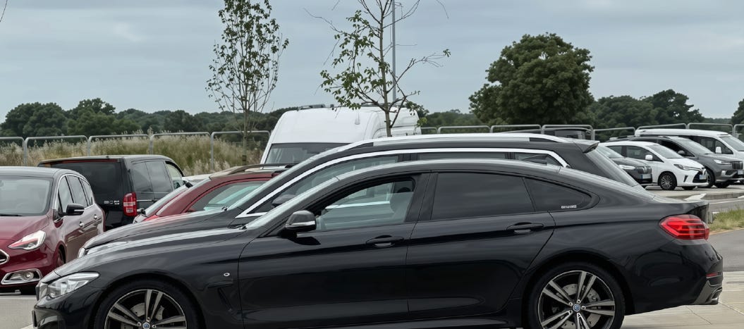 Under a cloudy sky and fields cordened off with Harras Fencing, cars are parked awaiting their children as its pick up time.  Many cars and vans parked along the new road, some blocking in others.