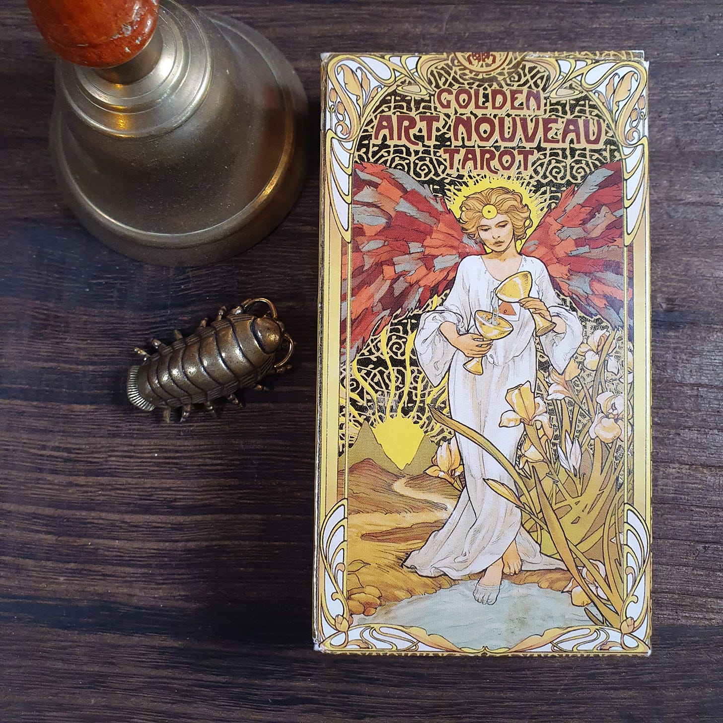Golden Art Nouveau Tarot deck box featuring Temperance angel illustration, placed beside brass bell and vintage insect object on wooden surface, photographed for the Tarot DMs interview with Alice Slater by Finbarre Snarey.