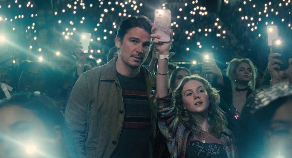 A white middle-aged man wearing a sweater and a jacket stands next to a white teenage girl holding up her phone with the flashlight on; they are surrounded by other audience members doing the same