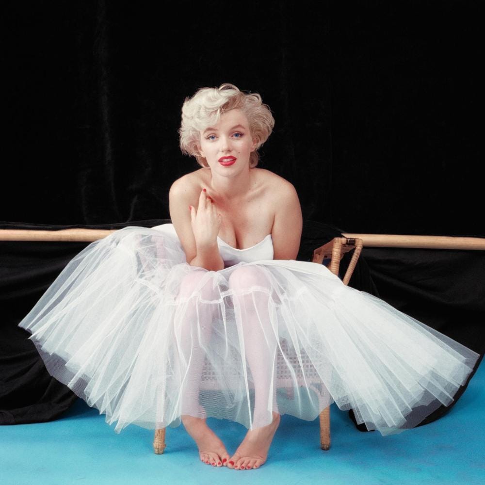 Colour full-length photo portrait of Marilyn Monroe seated on a chair wearing a white ballerina's tutu with bustier. She leans forward looking into the camera, her right hand raised to her collar bone. Colour full-length photo portrait of Marilyn Monroe seated on a chair wearing a white ballerina's tutu with bustier. She leans forward looking into the camera, her right hand raised to her collar bone.