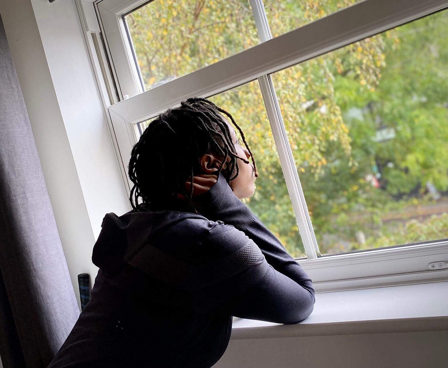 Sandra with short black locs, looks out the window with green leaves