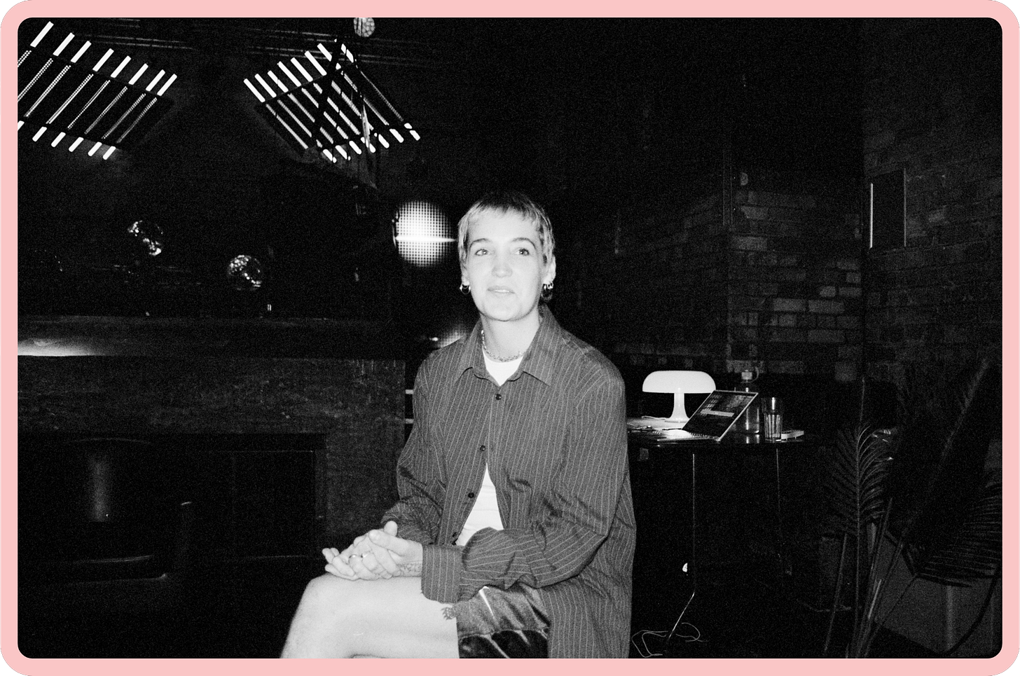 A black-and-white photo of Ali, a young woman with short blonde-brown hair. She is looking off-camera and wears a slight smile, and her hands are crossed in her lap. She is wearing a dark, striped oversized shirt and black shiny shorts.