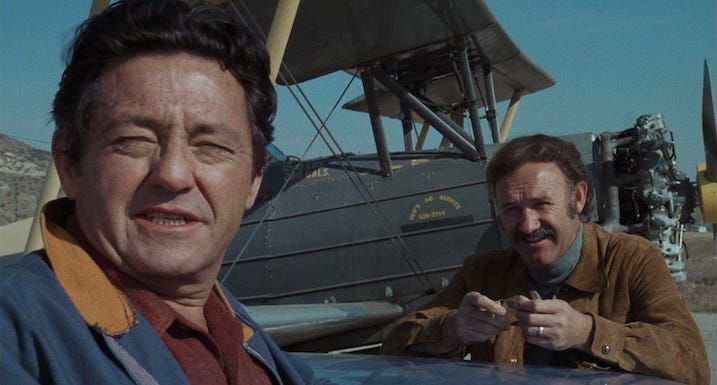 Gene Hackman smiles in the background while a man in a windbreaker speaks in front of a vintage biplane on a clear sunny day.