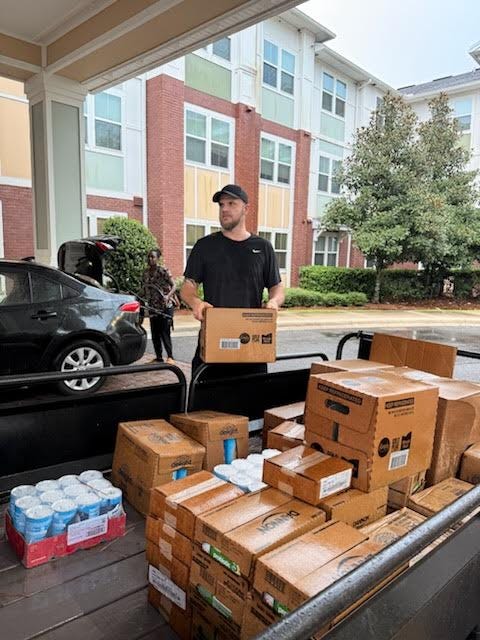 A man wearing a black shirt and black cap is unloading boxes from a trailer filled with cardboard boxes and canned beverages. Another person is visible in the background near an open car trunk. The scene takes place outdoors in front of a modern multi-story apartment building with large windows, red brick façade, and white trim. Trees and shrubs are also present near the building. No recognizable figures, landmarks, or brands are clearly identifiable. A man wearing a black shirt and black cap is unloading boxes from a trailer filled with cardboard boxes and canned beverages. Another person is visible in the background near an open car trunk. The scene takes place outdoors in front of a modern multi-story apartment building with large windows, red brick façade, and white trim. Trees and shrubs are also present near the building. No recognizable figures, landmarks, or brands are clearly identifiable.