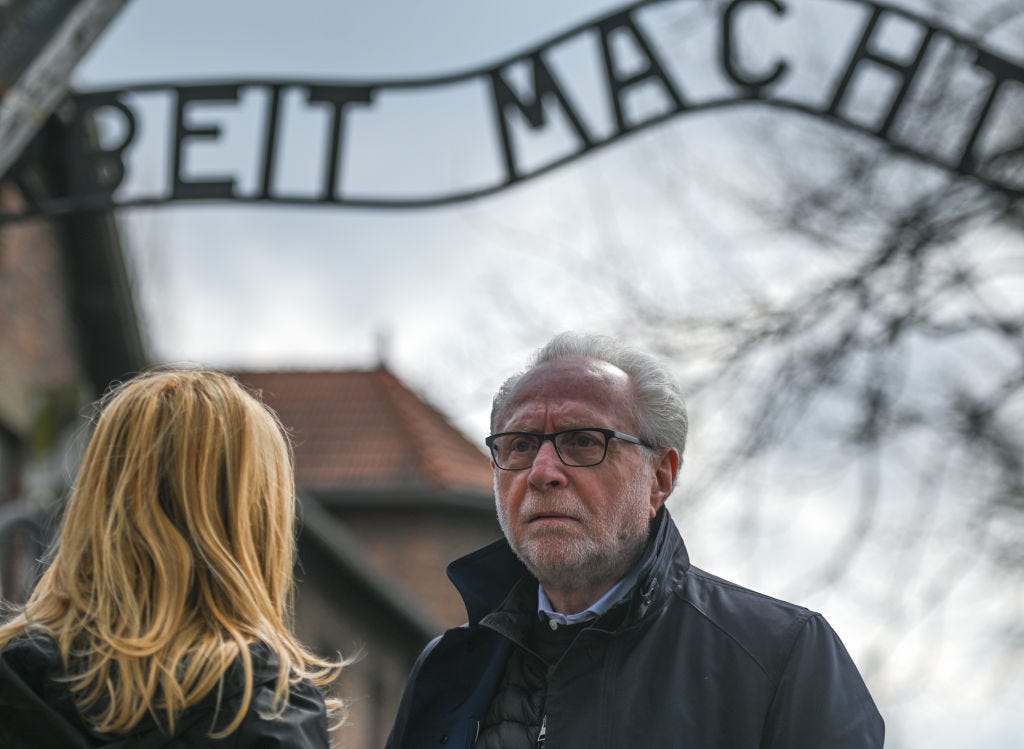 A man with white hair and close cropped beard in glasses and a dark coat stands under the sign to Auschwitz A man with white hair and close cropped beard in glasses and a dark coat stands under the sign to Auschwitz