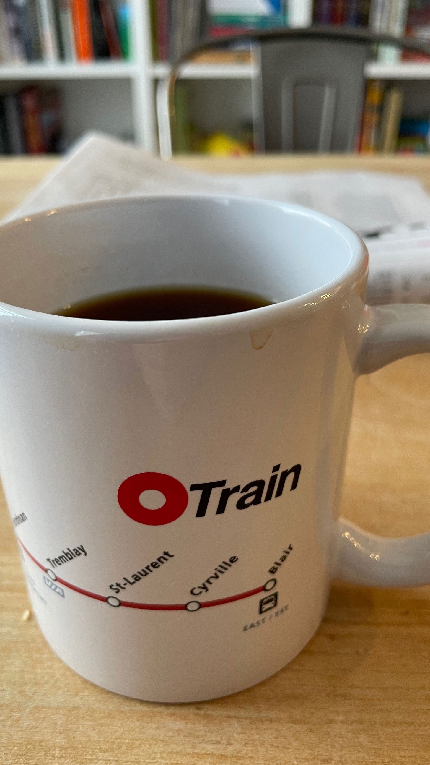 O-Train branded coffee mug with coffee in it. In the background, a newspaper and bookshelf out of focus O-Train branded coffee mug with coffee in it. In the background, a newspaper and bookshelf out of focus