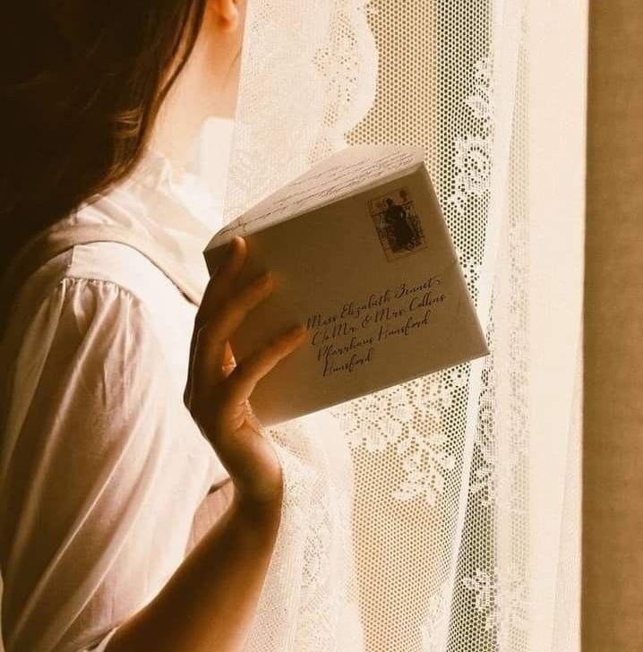 This may contain: a woman reading a book by a window with white lace on the curtain behind her