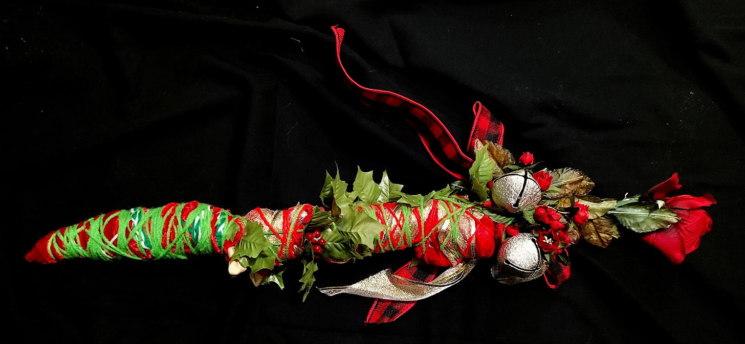 A decorated stick covered in red and green yarn with bells, bows, flowers and holly berries.