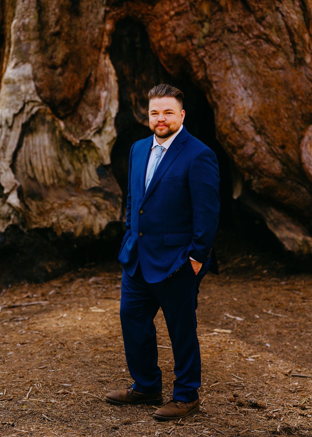 A full body picture of James wearing a blue suit and standing in front of a giant tree
