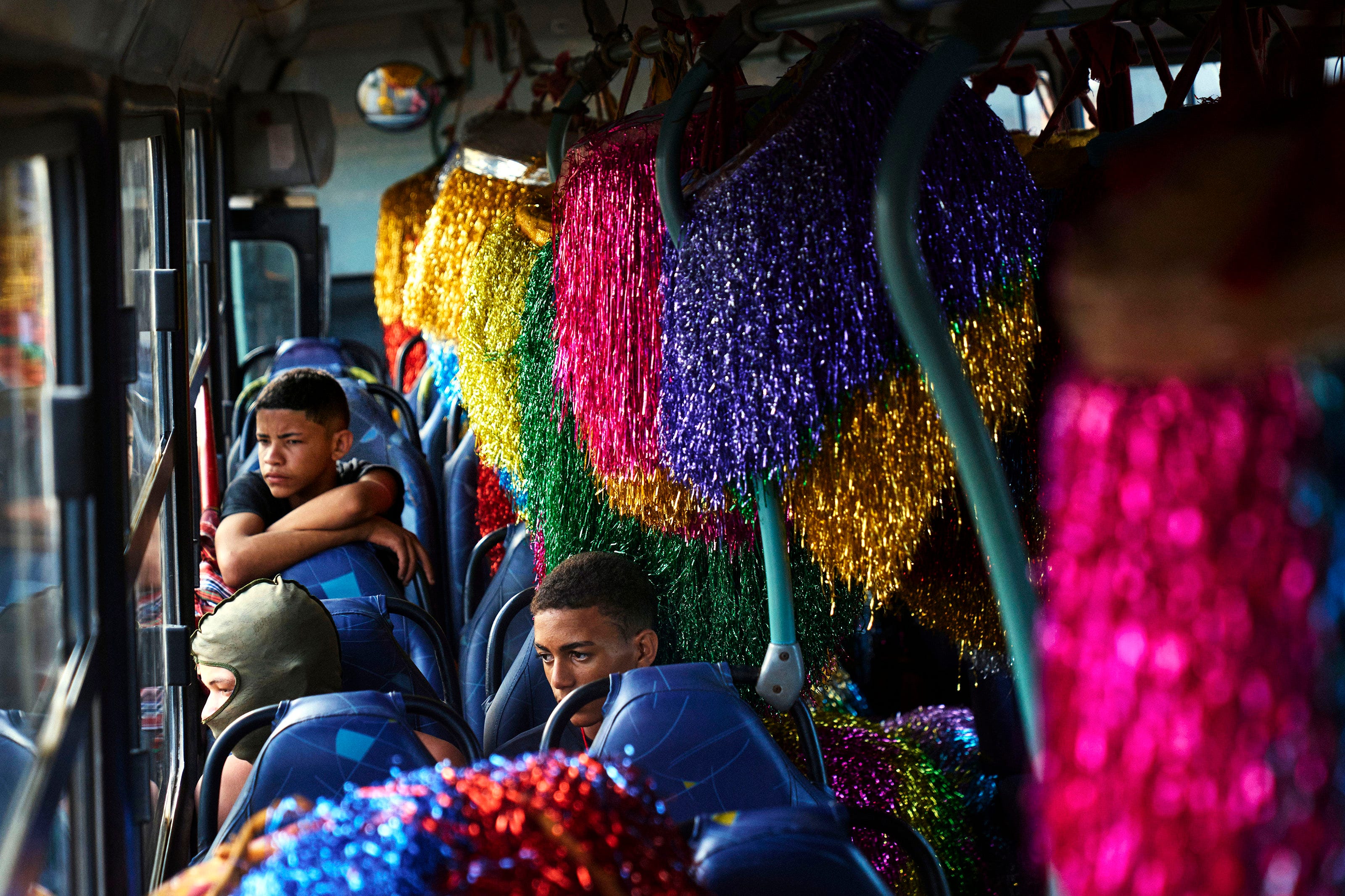 Several boys sit on a bus among colorful, shiny costumes with tinsel. One boy looks out the window while another wears a metallic mask, creating a festive, vibrant atmosphere inside the bus.