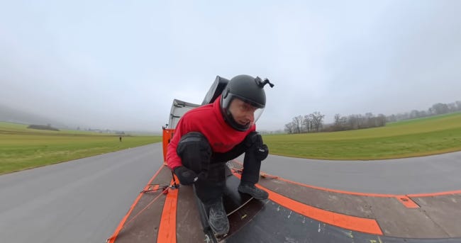 Le mécanisme de l'image du haut vu de plus près : sur une espèce de fauteuil pliant, un homme équipé d'un casque intégral à visière transparente fronce les sourcils. Le fauteuil semble monté sur des rails. Le mécanisme de l'image du haut vu de plus près : sur une espèce de fauteuil pliant, un homme équipé d'un casque intégral à visière transparente fronce les sourcils. Le fauteuil semble monté sur des rails.