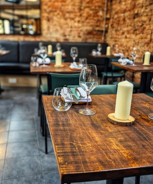 A square wooden table with glasses, plates, and a pillar candle on it, with other tables like this and a brick wall behind it