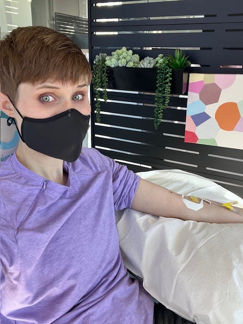 OP (a white person with a pixie cut and a purple shirt, wearing a grey face mask) sits in a white chair with an IV in her bared left arm, which is propped up on a pillow. Behind her, there's a black privacy divider with slats. To the left, there's a hanging "window" box of faux succulents and, to the right, a very small square print with blobs of abstract colors. OP (a white person with a pixie cut and a purple shirt, wearing a grey face mask) sits in a white chair with an IV in her bared left arm, which is propped up on a pillow. Behind her, there's a black privacy divider with slats. To the left, there's a hanging "window" box of faux succulents and, to the right, a very small square print with blobs of abstract colors.
