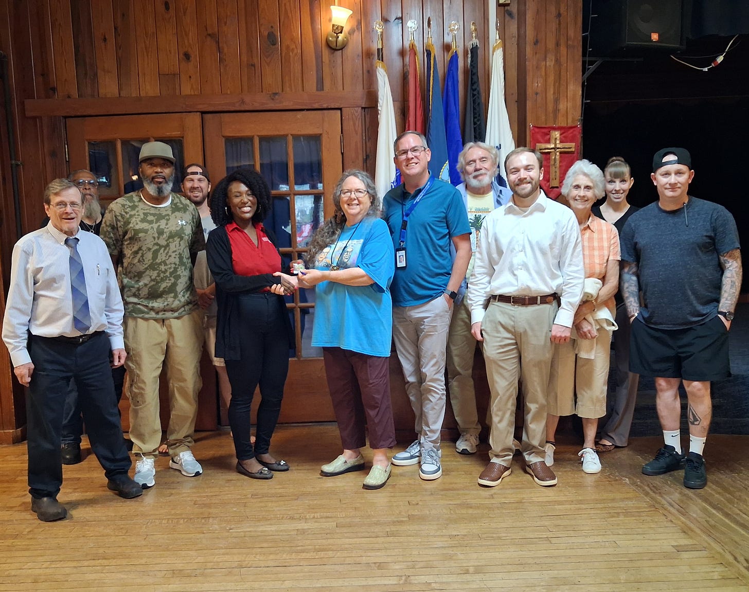 A group of people pose indoors in a wood-paneled hall with double doors behind them, several flags on stands, and a red banner with a gold cross. Two women in the center are shaking hands while holding a small item, and others smile around them; one man wears a camouflage Under Armour shirt, and another has visible arm tattoos and a backward cap. A group of people pose indoors in a wood-paneled hall with double doors behind them, several flags on stands, and a red banner with a gold cross. Two women in the center are shaking hands while holding a small item, and others smile around them; one man wears a camouflage Under Armour shirt, and another has visible arm tattoos and a backward cap.