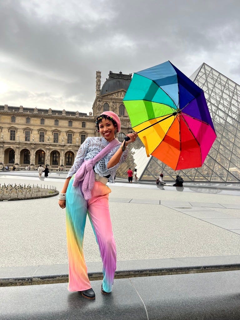 Kiana in a dressed by lala rainbow plisse suit holding a rainbow umbrella in front of the Louvre pyramids