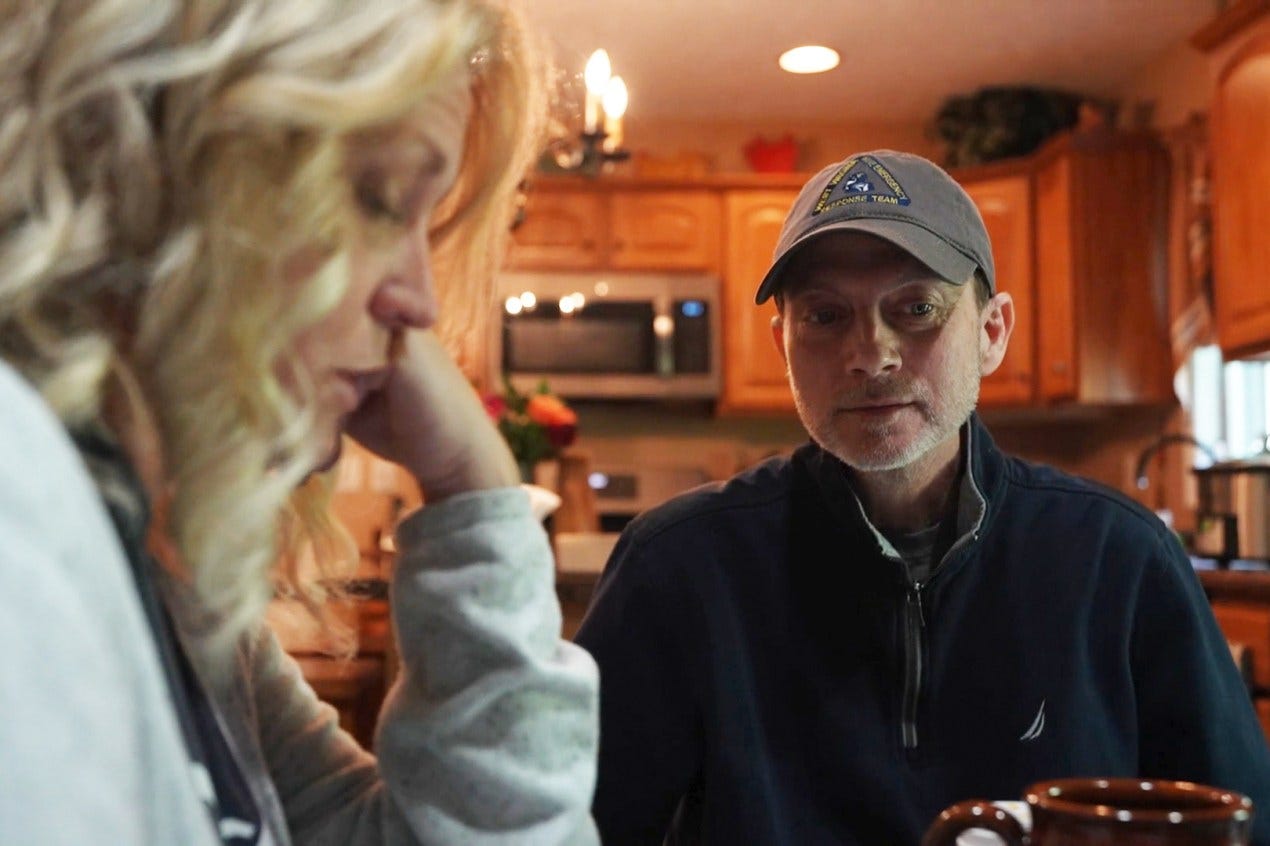 A husband sits at the kitchen table. His wife is seen close on the left of the frame. She is blurred slightly as the camera is focused on the husband. A husband sits at the kitchen table. His wife is seen close on the left of the frame. She is blurred slightly as the camera is focused on the husband.