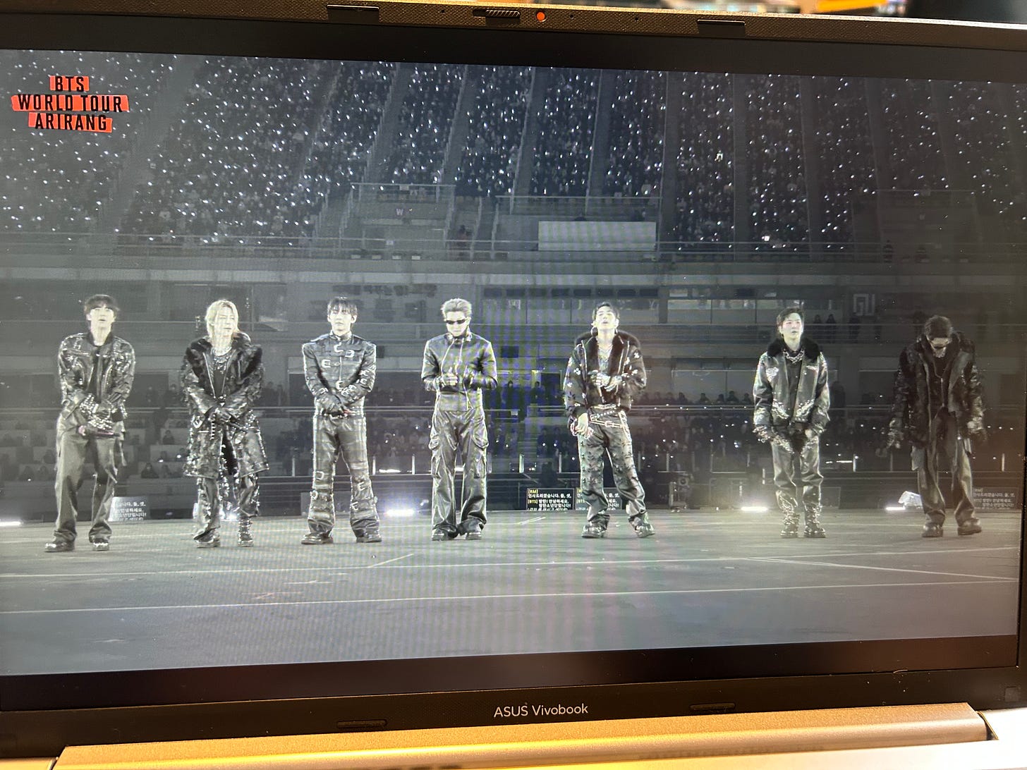The seven members of BTS in black leather performing on a stadium stage, seen through a laptop screen