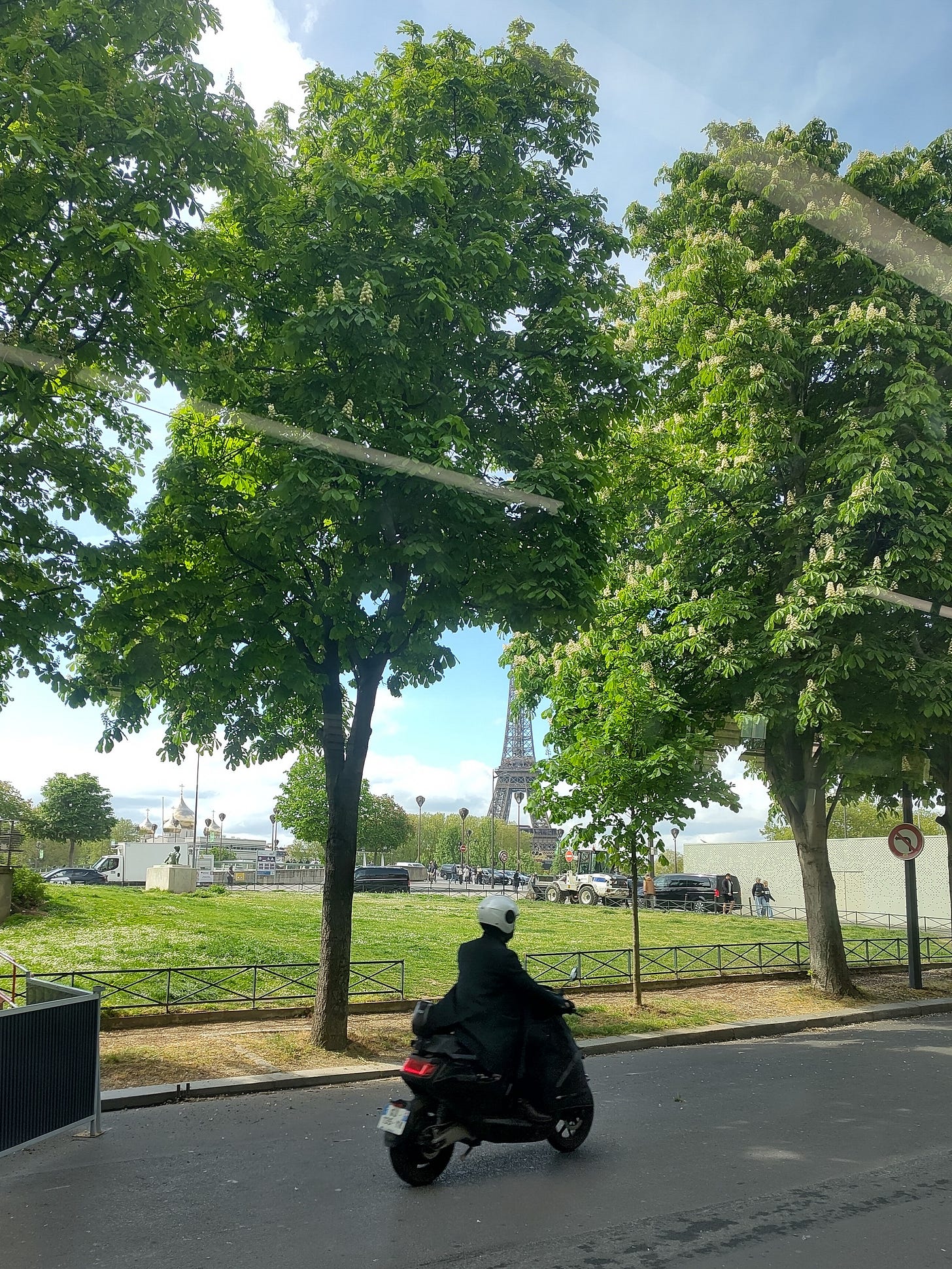 A person rides a motorcycle on the streets of Paris. In the background are lush green trees, a blue sky, and the Eiffel Tower.