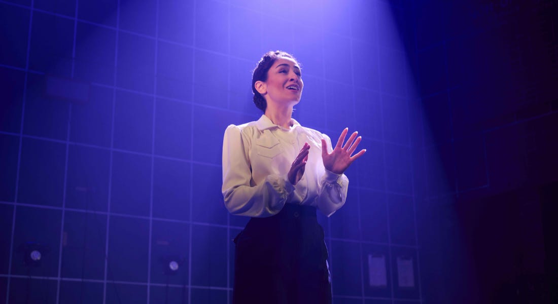 A woman on stage wearing a shirt and a black skirt, with a blue light background A woman on stage wearing a shirt and a black skirt, with a blue light background
