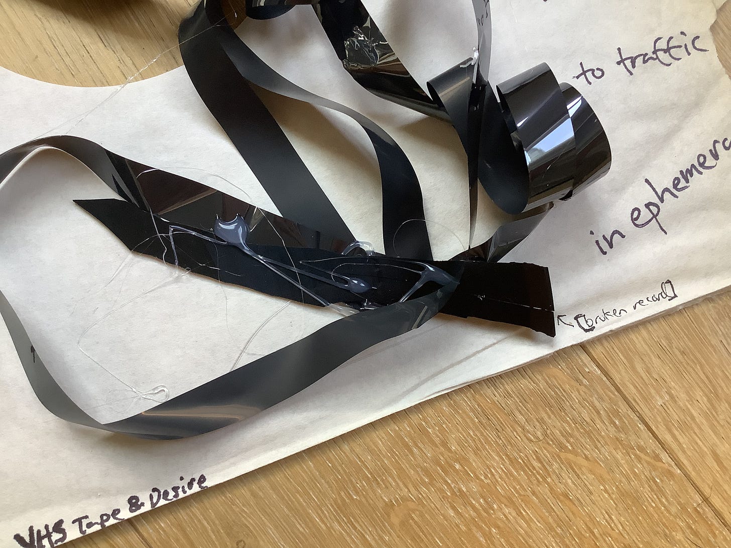 Detail with a large curl of magnetic tape glued together & an arrow nearby reading “[broken record]”, the text “to traffic / in ephemera” & “VHS Tape & Desire”