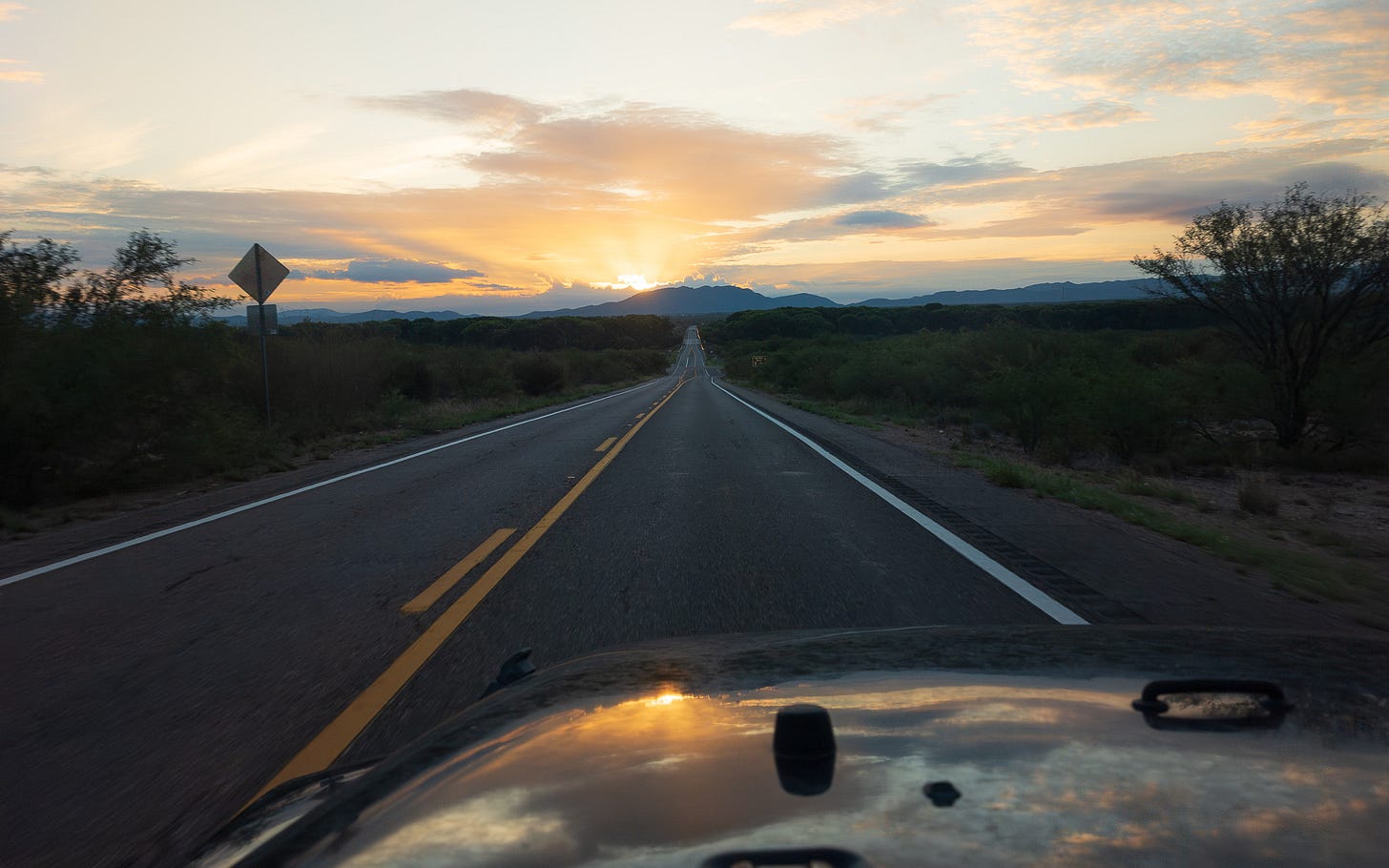 distant sunrise over a mountain, with a long road point toward it, and a reflection of the sky and colorful sunrise on the hood of the jeep the picture was taken from. distant sunrise over a mountain, with a long road point toward it, and a reflection of the sky and colorful sunrise on the hood of the jeep the picture was taken from.