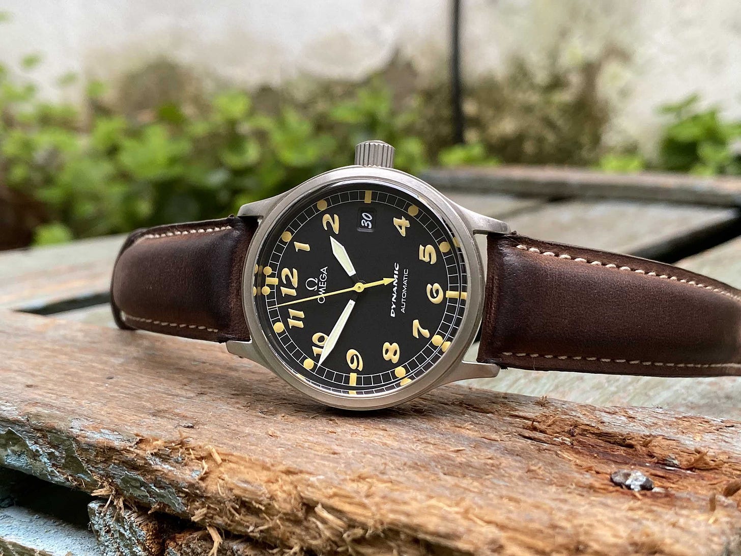 A close-up of an Omega Dynamic III watch on a wooden surface with greenery in the background.