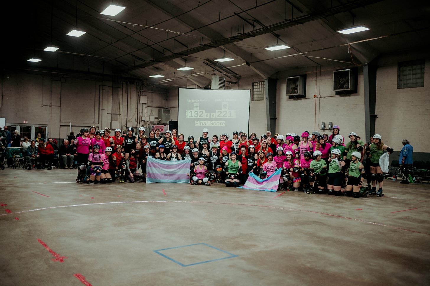 A large group of people, all dressed in different colored uniform tops and roller derby uniforms/safety equipment, pose holding two transgender pride flags. All of their shirts read "Trans Athletes Belong Here."
