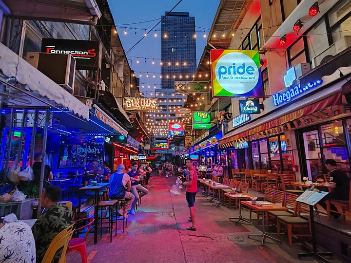 Silom 4 alley is full of neon signs, outdoor seating, and rainbow flags. A handsome queer Thai boy with a moustache poses in front of some erotic art at Go Grrrls