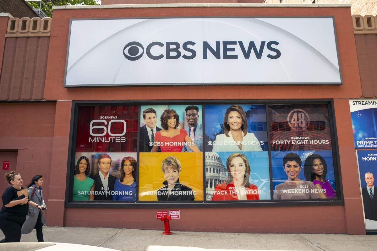 Two women walk past a sign for CBS news with photos of news anchors