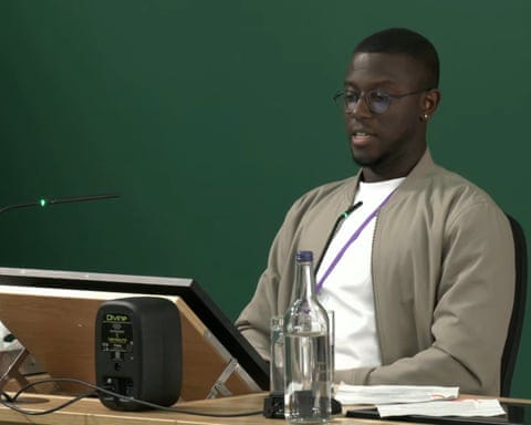 Elias Calocane in glasses and a beige jacket sits at a desk with a microphone, water bottle, and papers.