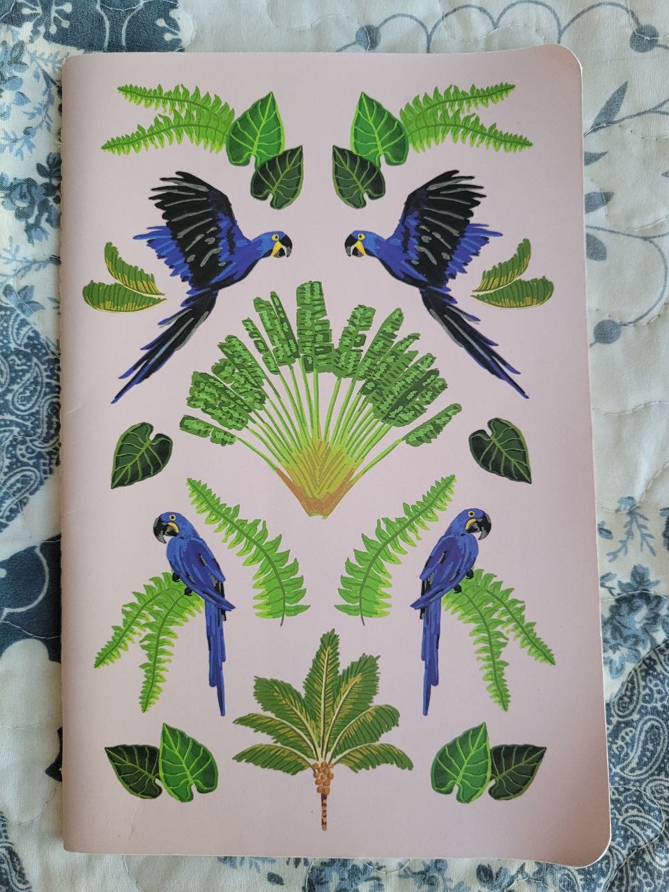 Caderno em cima de uma cama com colcha florida azul e branco. O caderno tem um tom rosado e desenhos de folhas e araras.