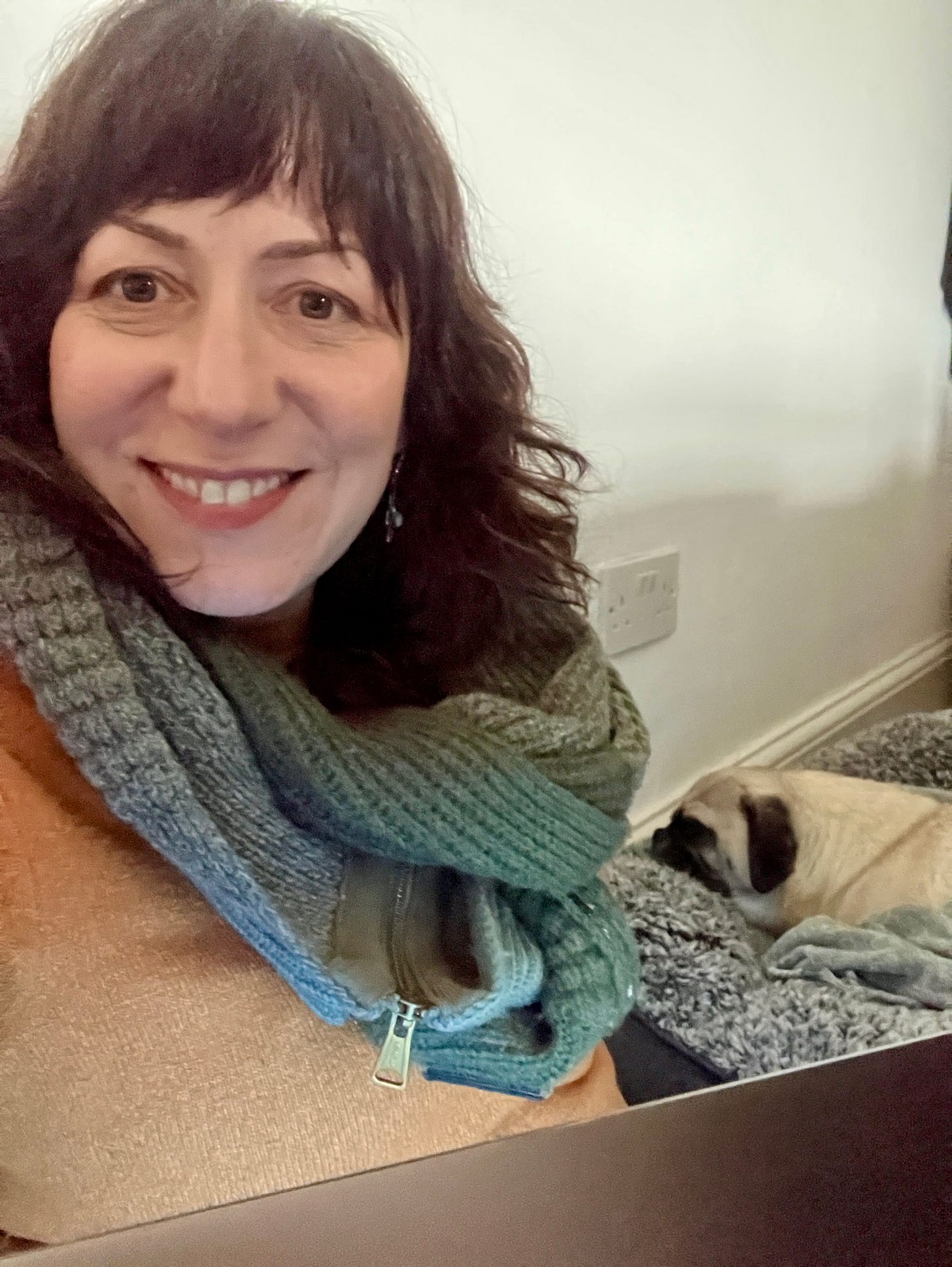 Kim is sitting on the floor with her laptop taking a selfie. Terry the Pug sleeps in the background.