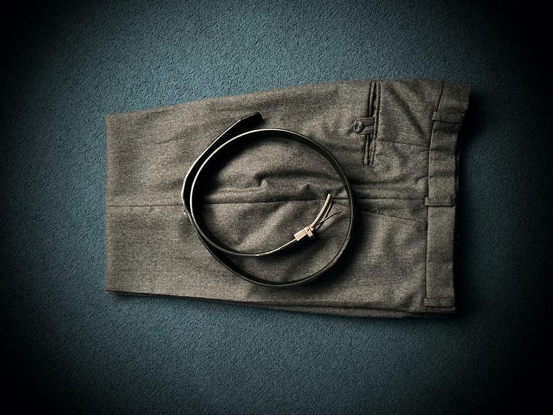 A pair of grey trousers, folded in half with a belt coiled up and placed on them.