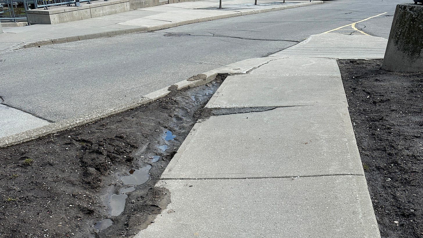 A sidewalk at Jackson Street West and Bay Street South at the western entrance to City Hall showing visible cracking, asphalt repair, and tilting A sidewalk at Jackson Street West and Bay Street South at the western entrance to City Hall showing visible cracking, asphalt repair, and tilting