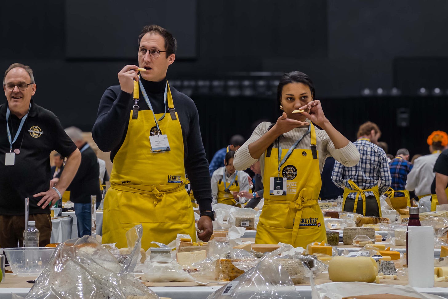Jury-Mitglieder der World Cheese Awards 2025 in Bern stehen an einem grossen Verkostungstisch und beurteilen Käsestücke. Im Vordergrund stehen eine Frau und ein Mann in gelben Schürzen, beide riechen oder schmecken gerade an einem kleinen Stück Käse. Der Tisch ist dicht gefüllt mit Laiben, Proben und Verpackungen. Im Hintergrund arbeiten weitere Personen konzentriert an ihren Tischen.