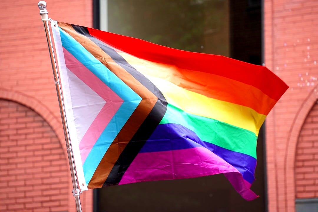 A rainbow colored flag on a pole in front of a building