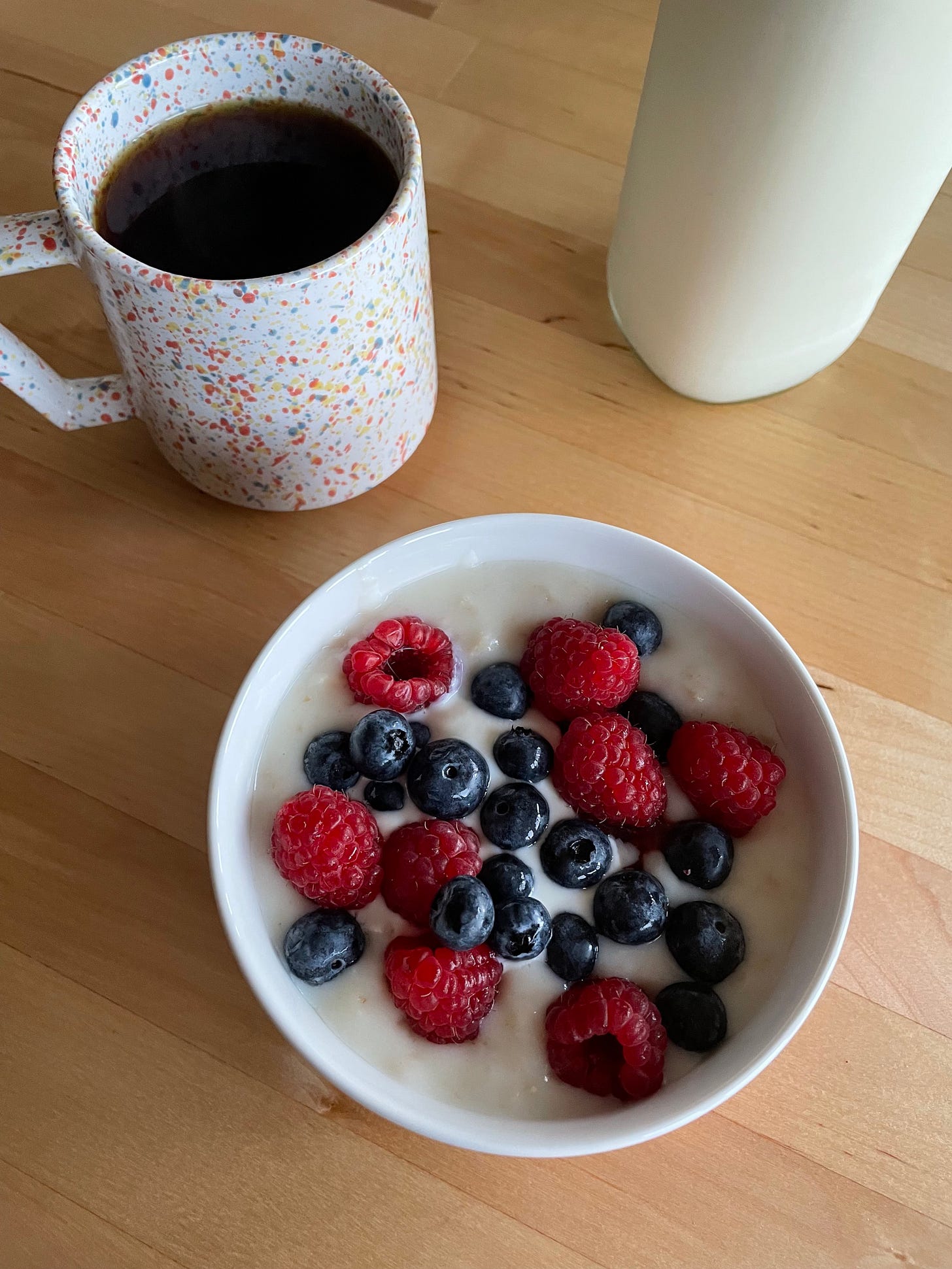Mug of coffee, bowl of porridge topped with berries, a bottle of milk in the background.