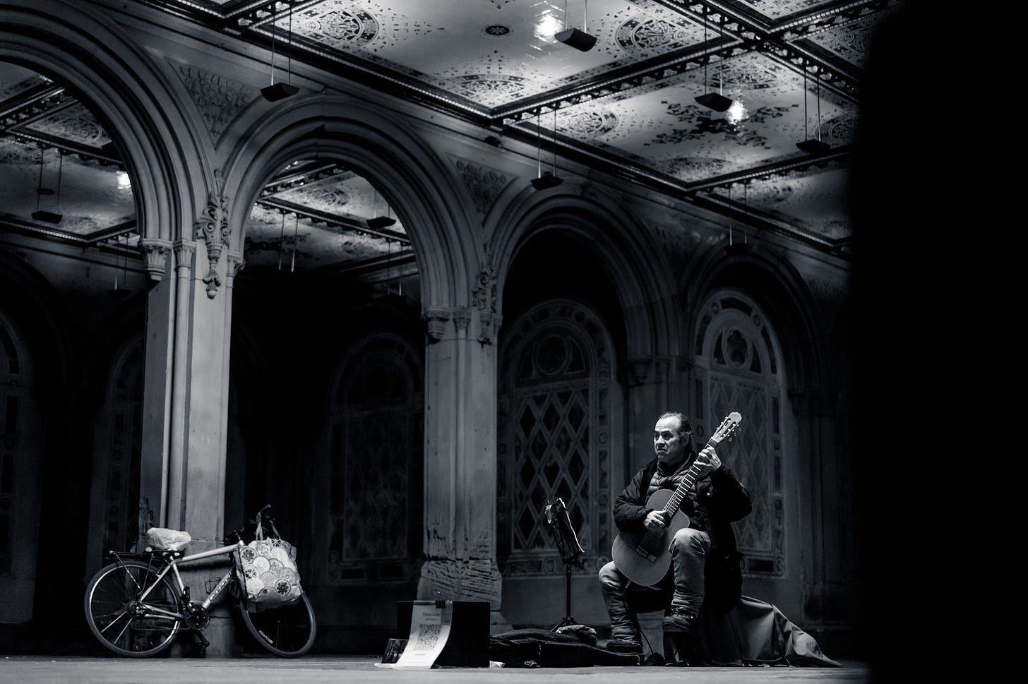 Classical guitarist Carlos König seated playing beneath the tiled arcade ceiling of Bethesda Terrace in Central Park, a bicycle propped against a column, New York, in black and white.