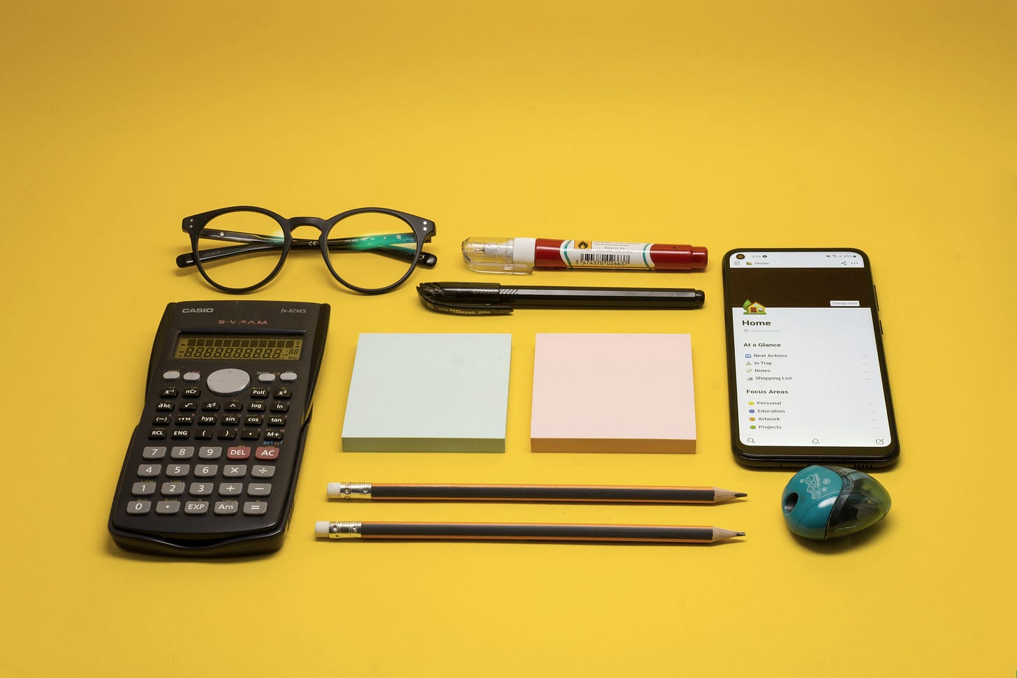 On a smooth yellow surface the following items are neatly arranged: black rimmed reading glasses; two pens; one black calculator; two post it blocks, one blue and one pink; two sharpened pencils; one pencil sharpener; a smartphone showing a list under the title “Home”.
