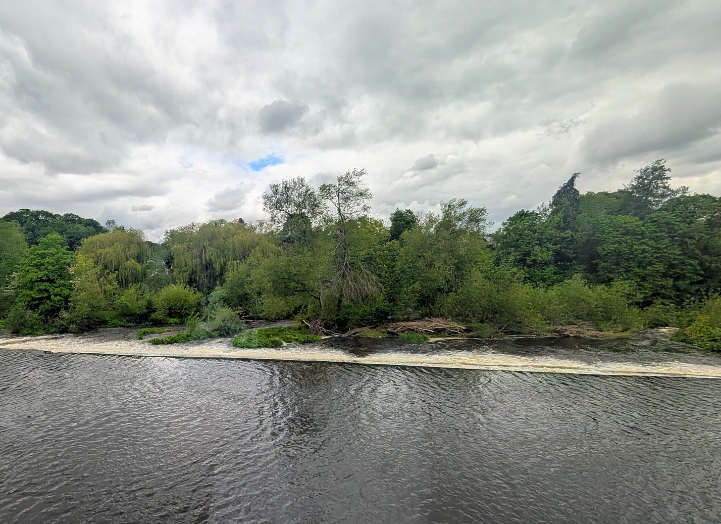 View through a restaurant window onto the River Wharfe in Otley, West Yorkshire. The river is running fast with water roaring over a weir. Trees line the far bank and a heavy, clouded sky fills the upper half of the image.