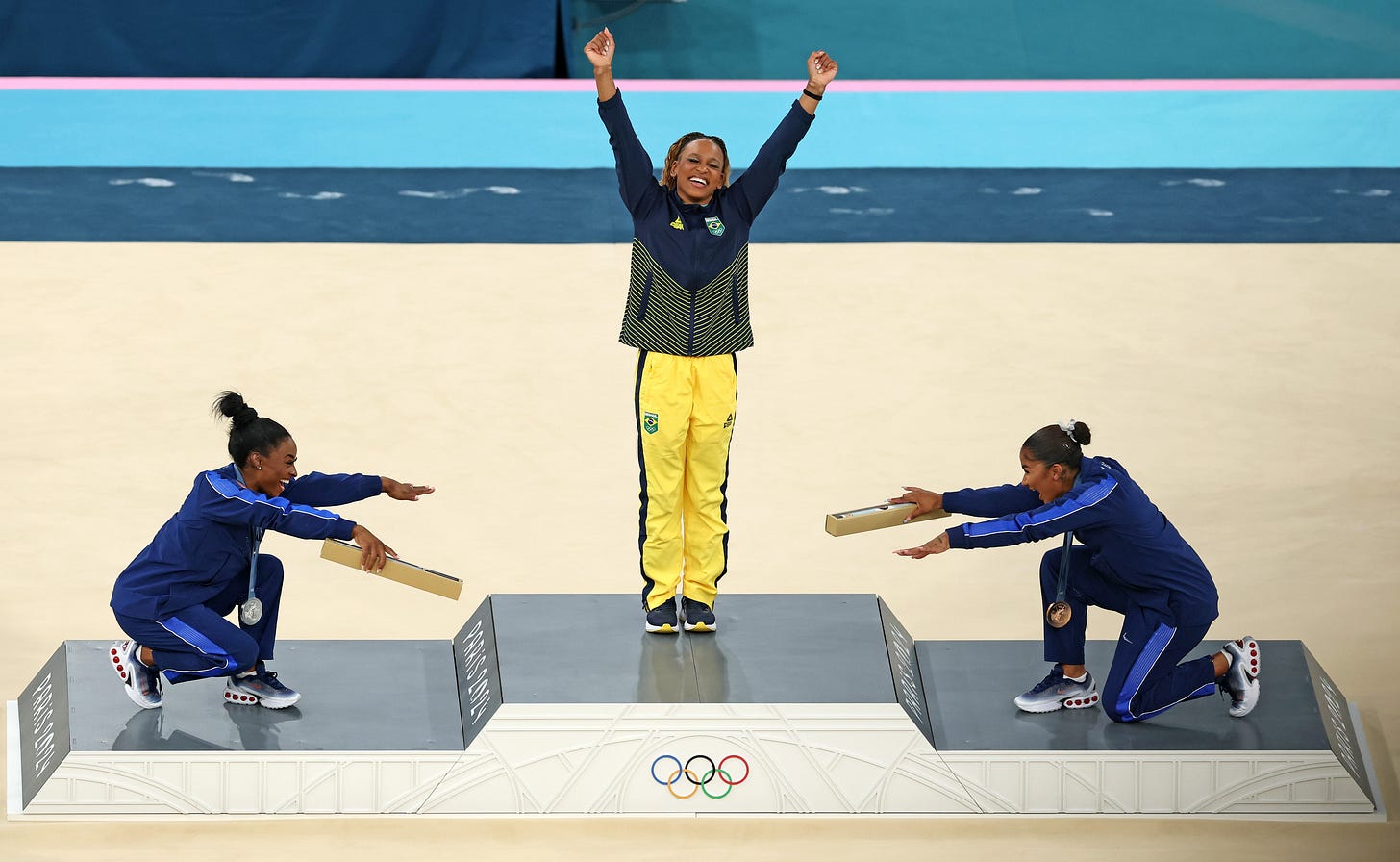 On a medals podium, two gymnasts on either side of the gold medal winner playfully bow down to her, as she raises her arms. On a medals podium, two gymnasts on either side of the gold medal winner playfully bow down to her, as she raises her arms.