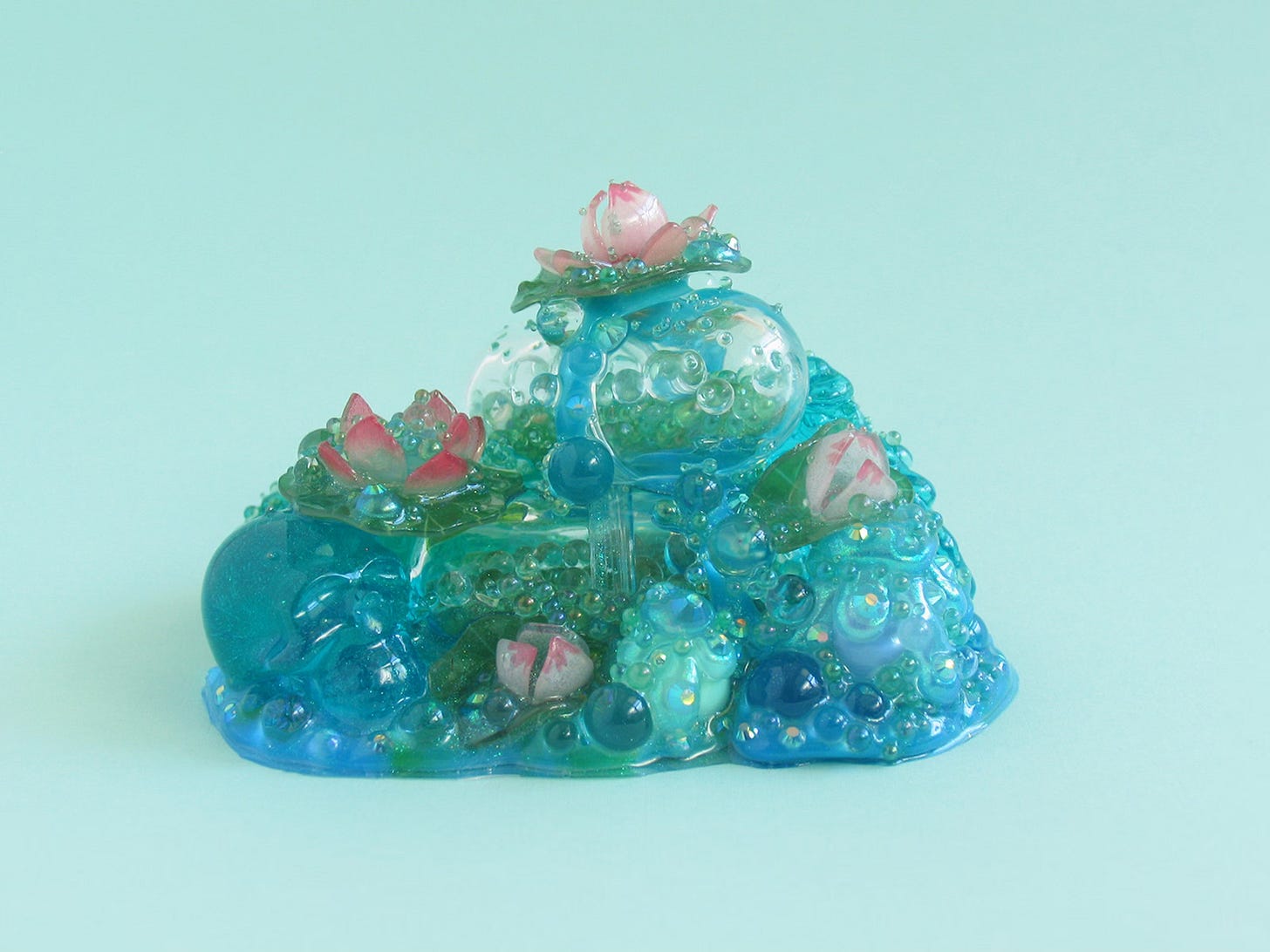 A miniature Glue Baby sculpture: pills full of dark green beads stacked on top of melty blue base with a blue skull on a left; lotus flowers top the pills, off of which is on a mint green background