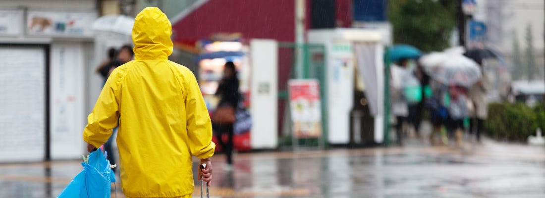 A person in a yellow rain jacket with their back to the camera, they are facing other people off in the distance who are beside some vending machines and holding umbrellas. A person in a yellow rain jacket with their back to the camera, they are facing other people off in the distance who are beside some vending machines and holding umbrellas.