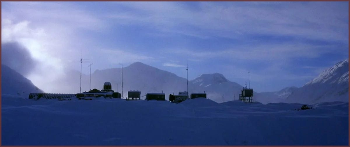Film frame from The Thing (1982): wide exterior shot of Outpost 31 at dusk, the compound's low grey structures and radio antennas silhouetted against snow-covered mountains. Shot on location at Salmon Glacier, Stewart, British Columbia.