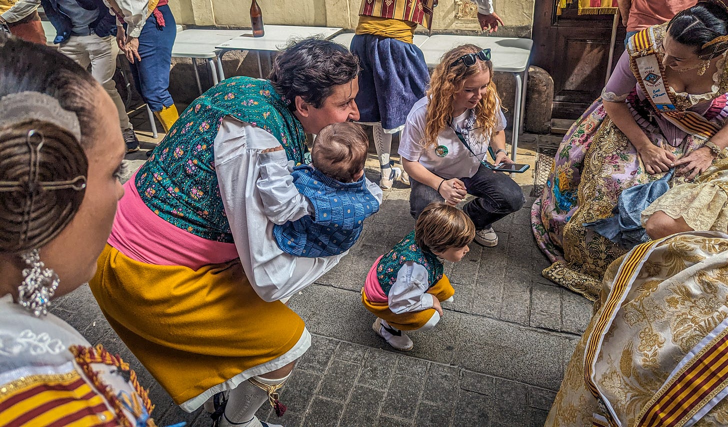 A father wearing traditional clothing holds his infant son in his arms as they participate in the pasodoble. His four old son is next to him. 