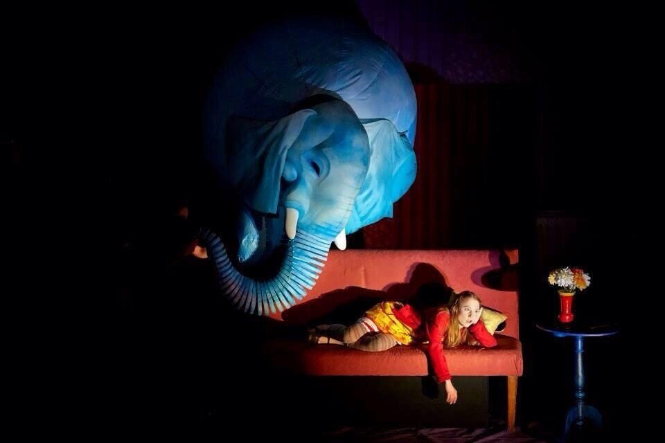 Small woman reclines on a sofa on a stage. Behind her is a massive, blue, beautiful elephant puppet.