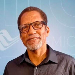 Homem negro de meia-idade, com cabelos curtos e grisalhos e barba curta e grisalha. Usa óculos de armação escura e veste uma camisa cinza. Fundo, parede azul clara com desenhos abstratos.