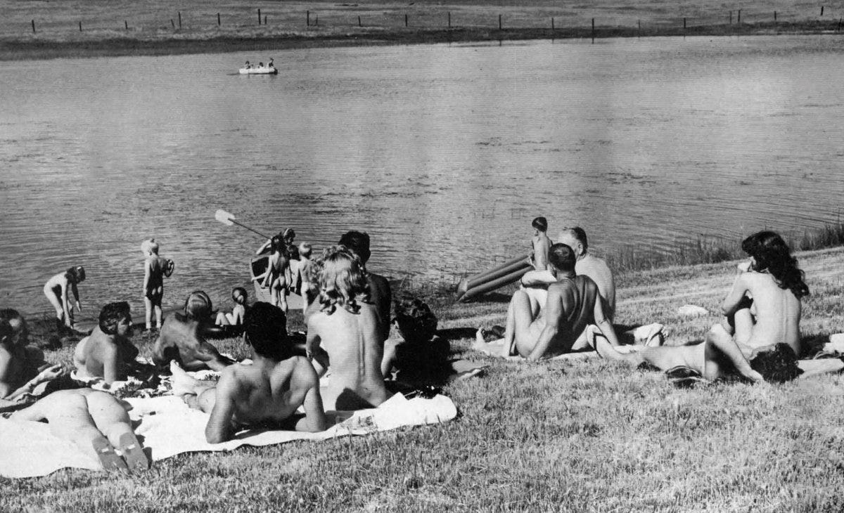Black and white photo of nude adults and children relaxing on a grassy lakeshore at Laguna del Sol, with swimmers and a small boat visible in the water.