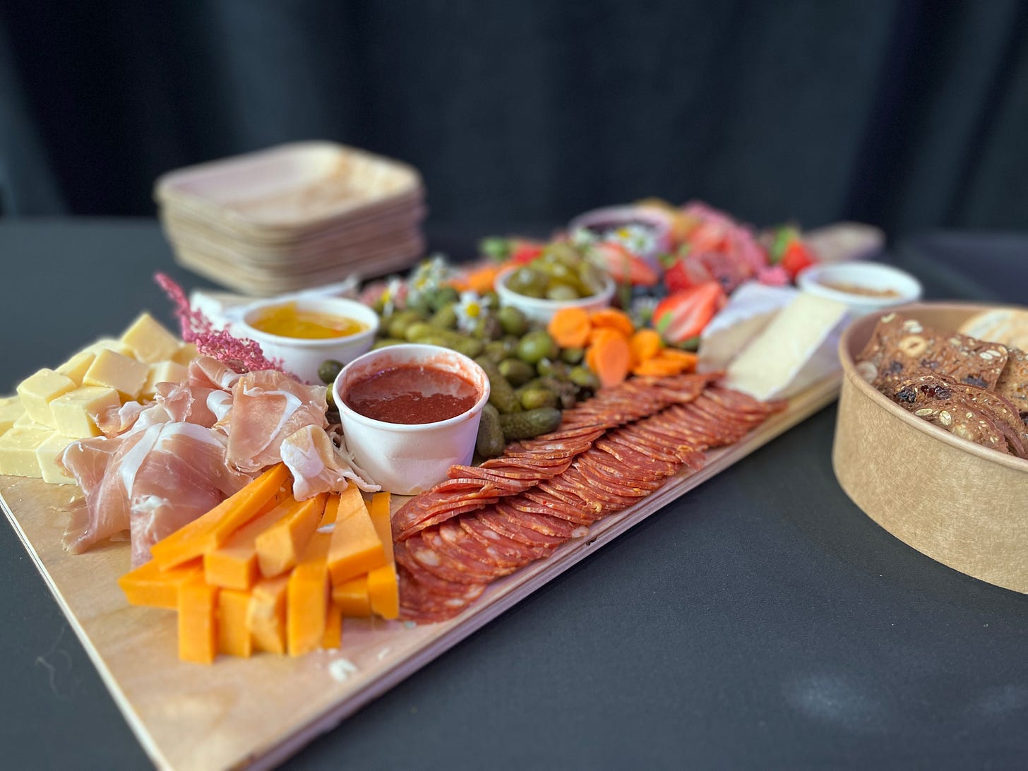 Charcuterie with a selection of sliced deli meat, cheese, veggies and dipping sauces. A bowl of crackers sits off to one side. Charcuterie with a selection of sliced deli meat, cheese, veggies and dipping sauces. A bowl of crackers sits off to one side.