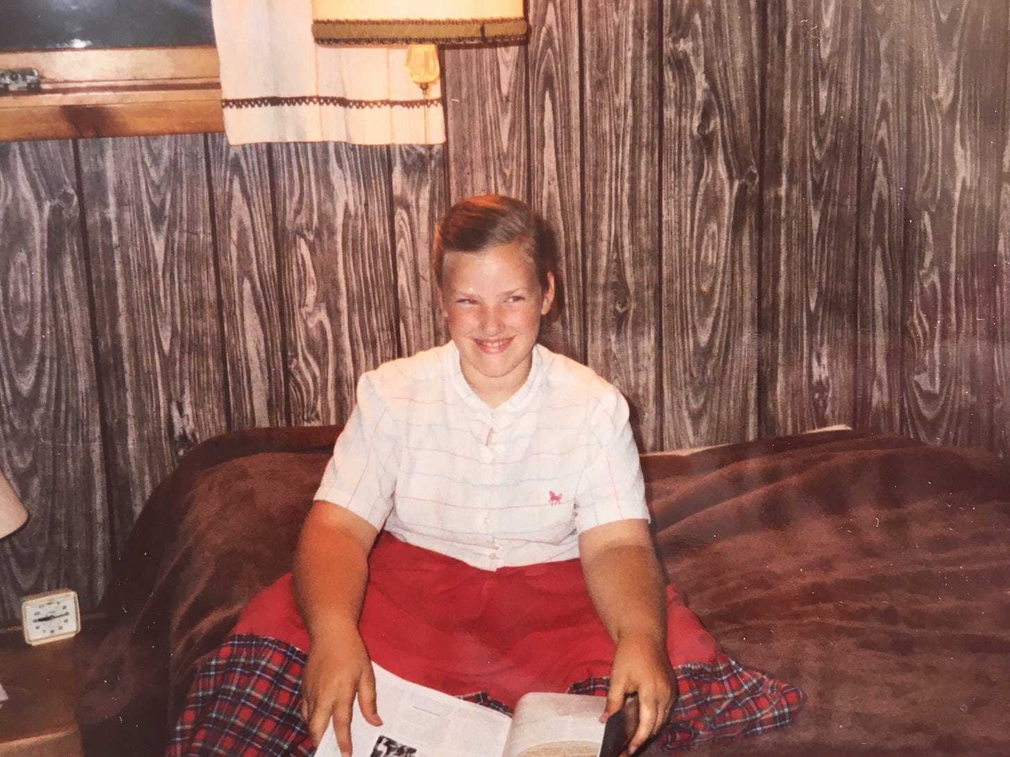 A young white girl with light brown hair pulled back in a braid sits cross-legged on a double bed. She’s wearing a white button down shirt with blue and pink stripes and a long red skirt with a wide black and red plaid ruffle on the bottom. She’s holding a magazine in her hands resting on her lap and looking over at someone while smiling. 